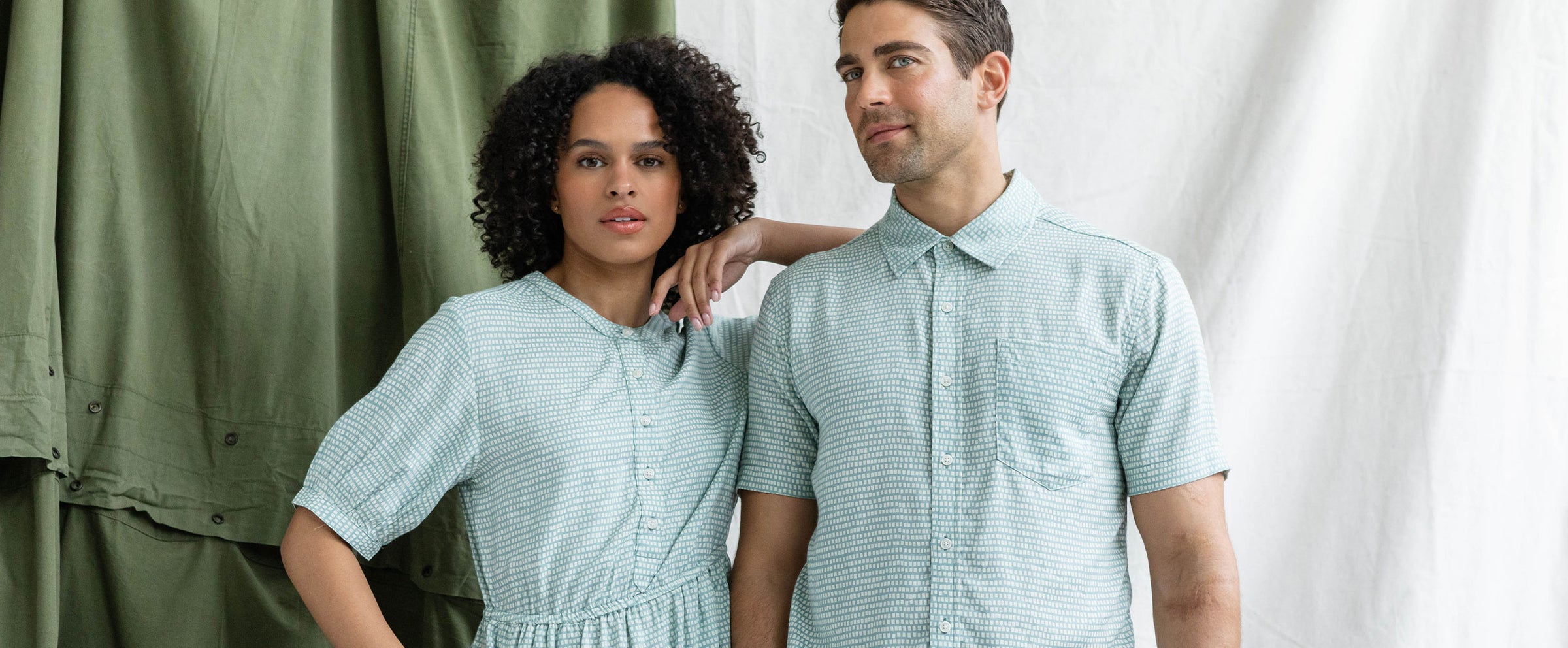 A woman and a man stand side by side wearing light blue and white striped outfits, posing in front of green and white draped fabric. The woman rests her arm on the mans shoulder.