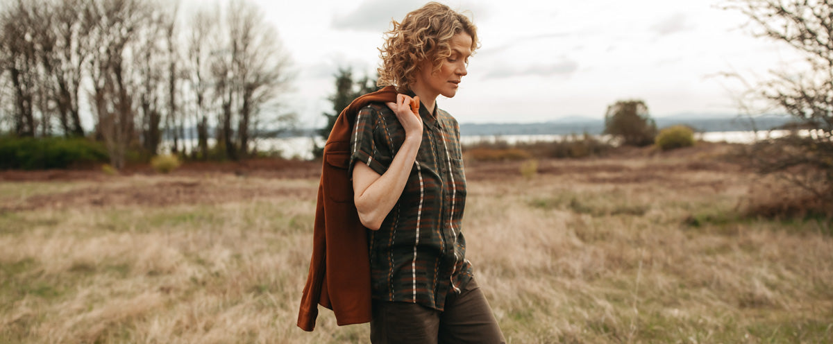 A person with curly hair walks through a grassy field, holding a brown jacket over their shoulder. The landscape is open with trees, bushes, and a body of water in the background under a cloudy sky.