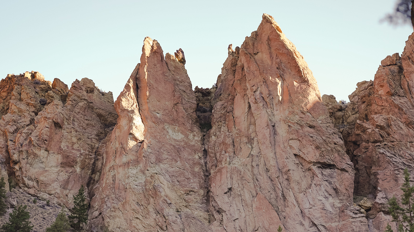 Jagged, sunlit rock formations rise vertically against a clear sky, with sparse vegetation at the base and a few trees visible in the lower corners of the image.