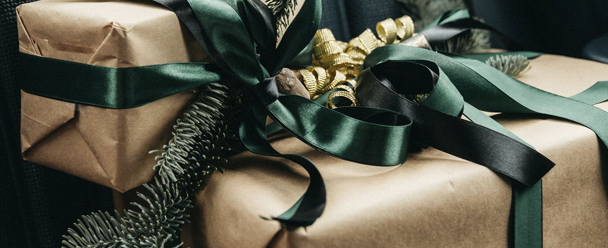 Two gifts wrapped in brown paper with dark green satin ribbons are adorned with pine branches and festive gold decorations. The scene evokes a cozy, holiday atmosphere.