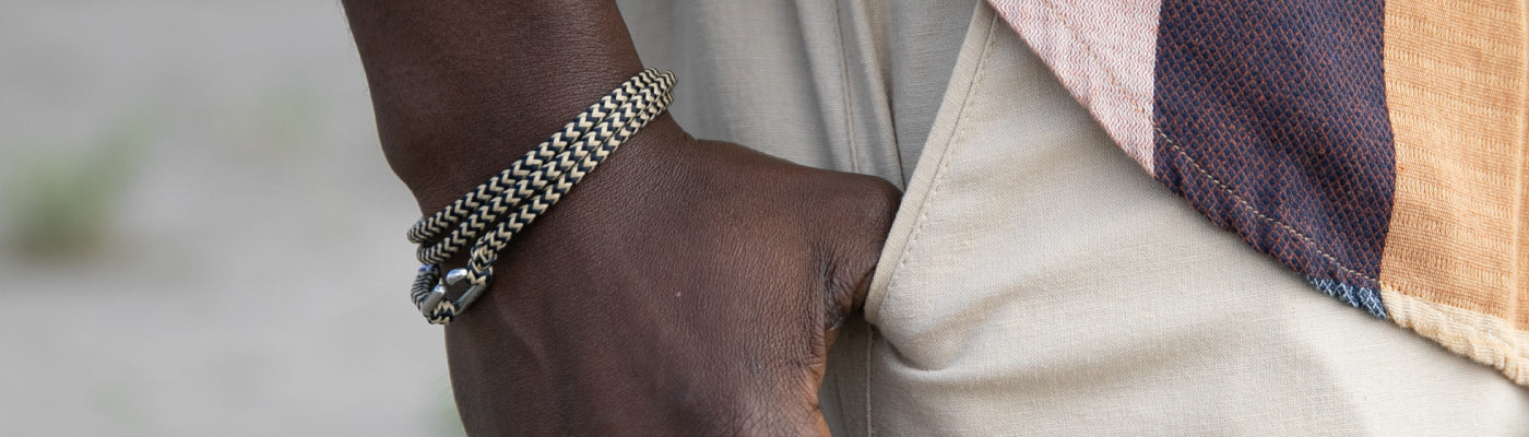 A close-up of a person’s hand wearing two braided bracelets, with their thumb hooked into the pocket of light-colored pants. The person is also wearing a textured, multicolored shirt.