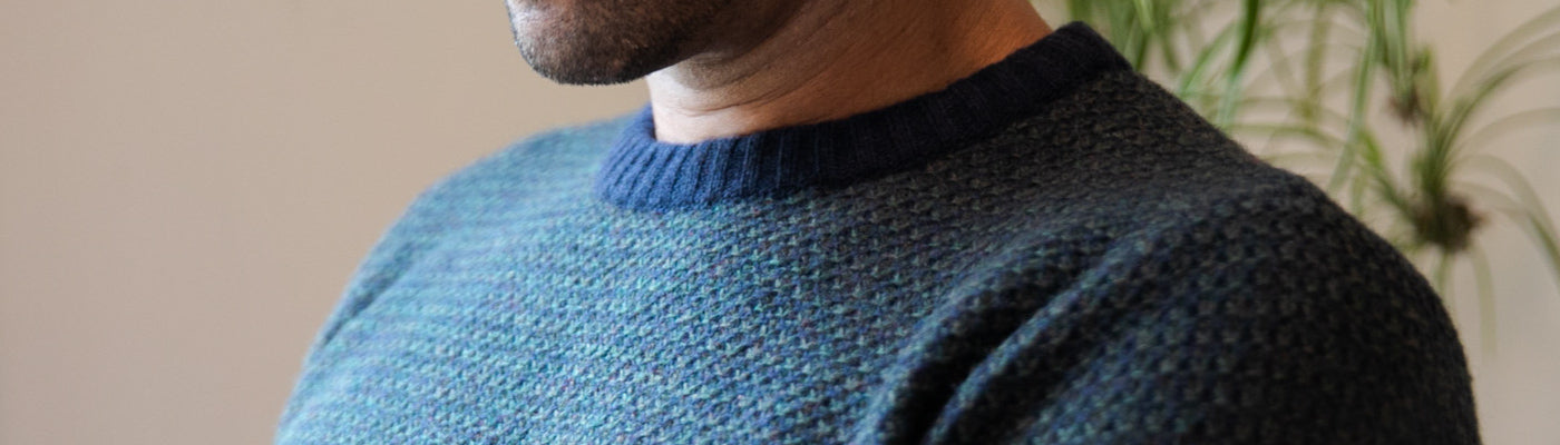 A person wearing a textured blue-green sweater with a navy collar, shown from the chin to chest. A blurred green plant is visible in the background.