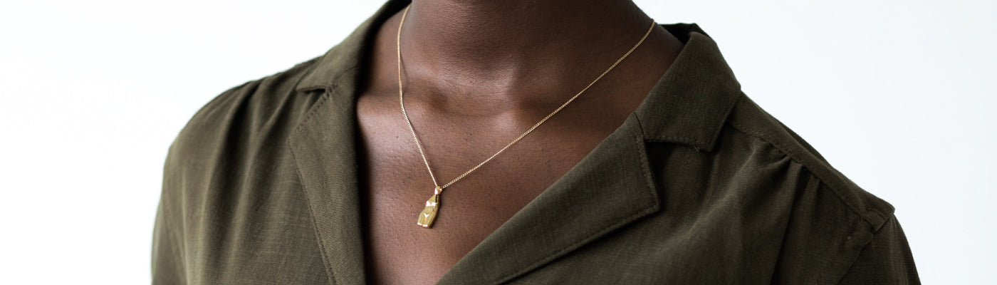 Close-up of a person wearing an olive green collared shirt and a delicate gold necklace with a rectangular pendant. The background is plain white.