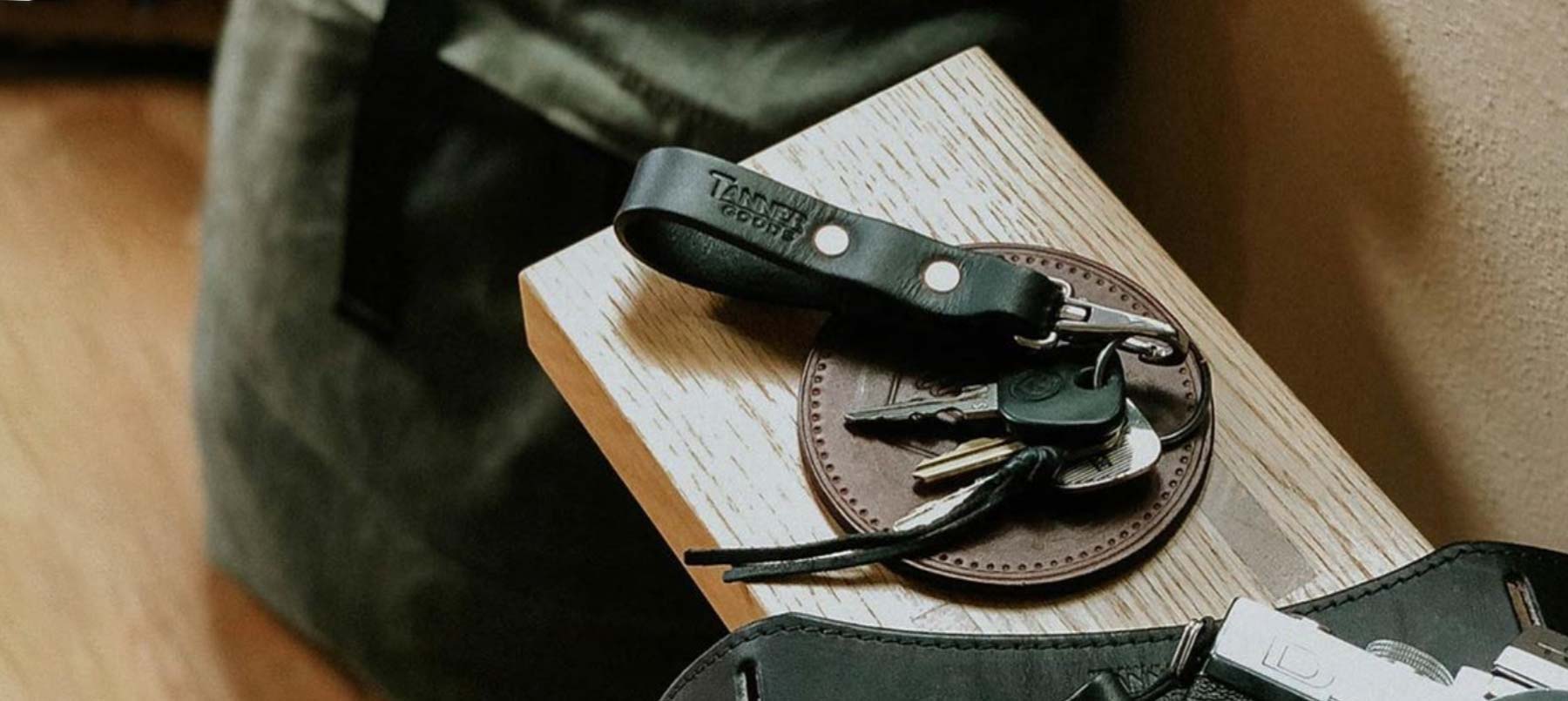 A set of keys with a black leather keychain rests on a round brown leather coaster, placed on a wooden surface next to a green bag.
