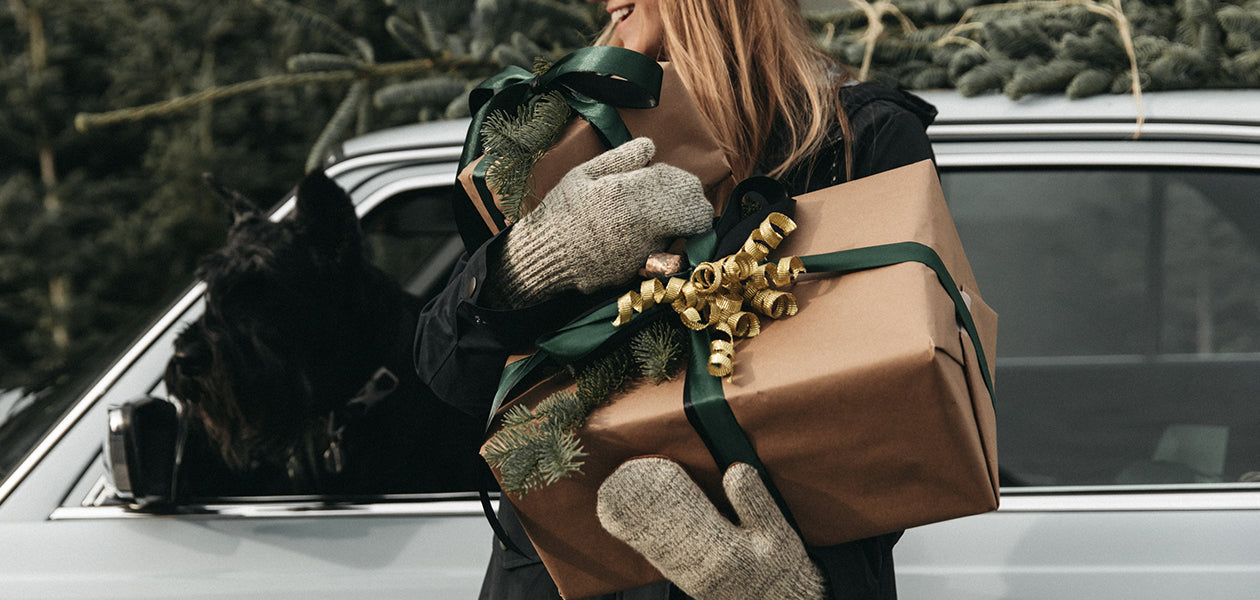 A woman wearing mittens carries gift boxes wrapped in brown paper with green ribbons, standing next to a car with a dog looking out of the window.