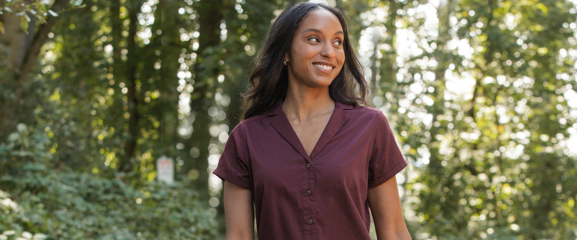 A woman in a short-sleeved maroon shirt stands outdoors, smiling and looking to the side. She is surrounded by greenery and trees with sunlight filtering through the leaves.