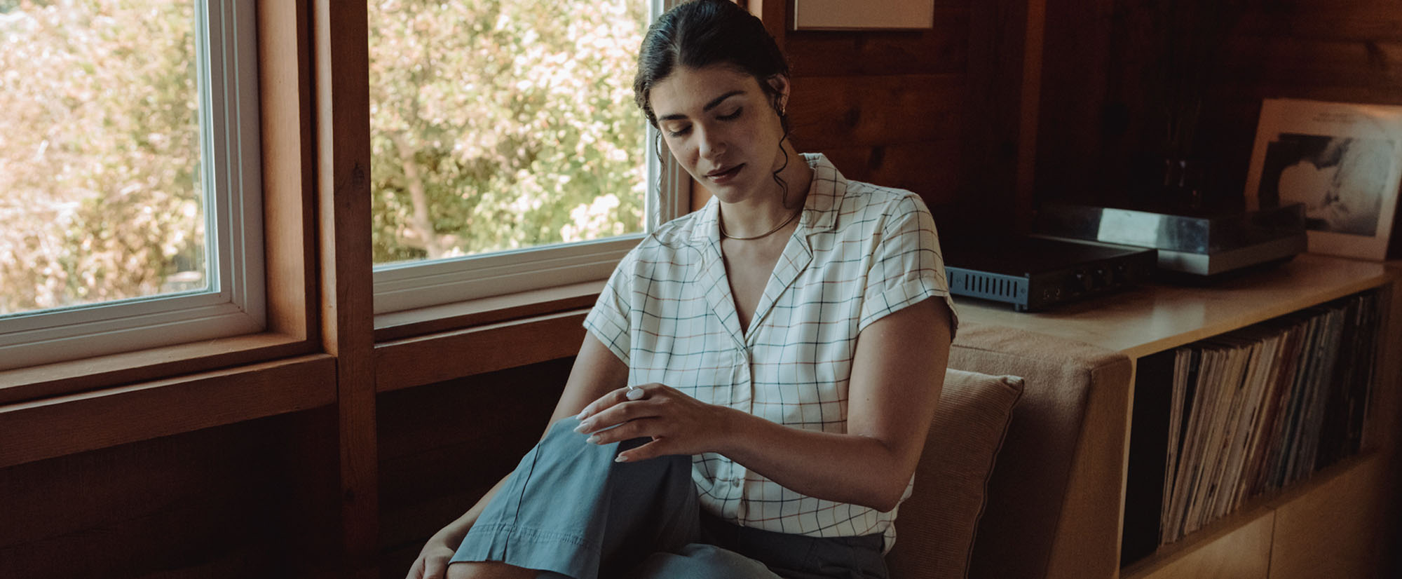 A woman sits indoors near a window with sunlight coming in, looking down thoughtfully. She wears a white plaid shirt and blue pants, with wooden shelves and records behind her.