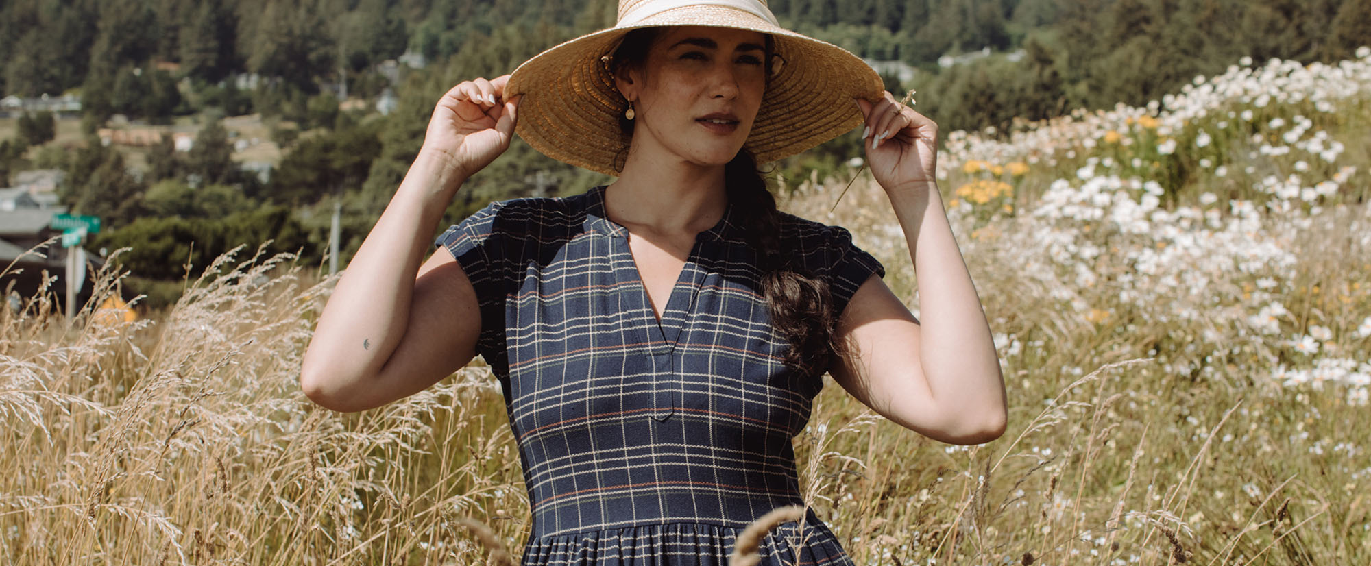 Woman wearing a straw hat and plaid dress stands in a field of tall grass and wildflowers, with trees and hills in the background on a sunny day.
