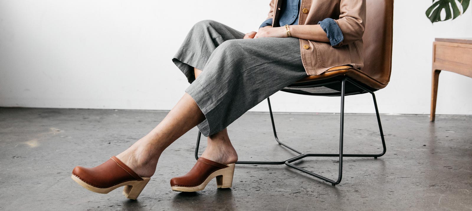 Person sitting on a modern chair wearing brown clogs, gray wide-leg pants, and a tan jacket with rolled-up sleeves. The setting is a minimalist room with a concrete floor and wooden furniture.