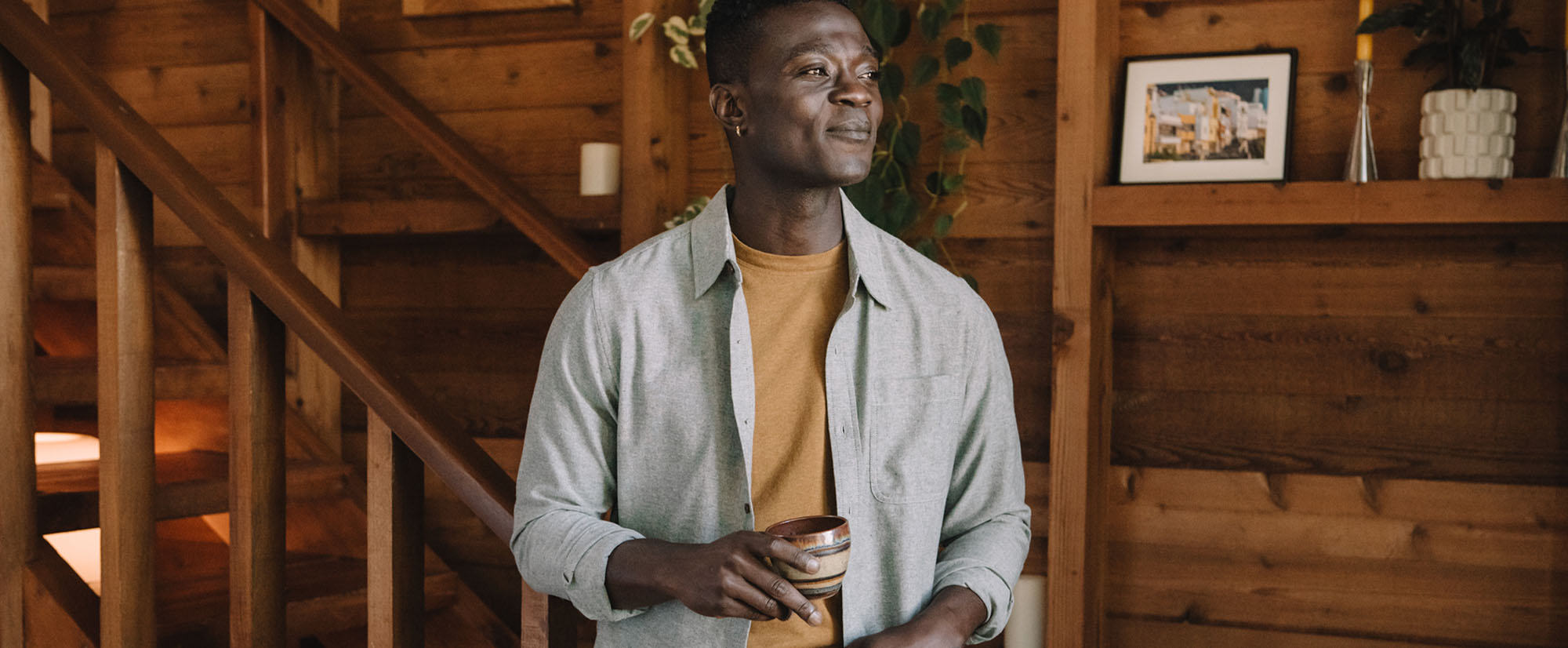 A man holding a mug stands in a cozy wooden room near a staircase, wearing a light shirt over a mustard t-shirt, with plants, a photo, and decor on the walls behind him.
