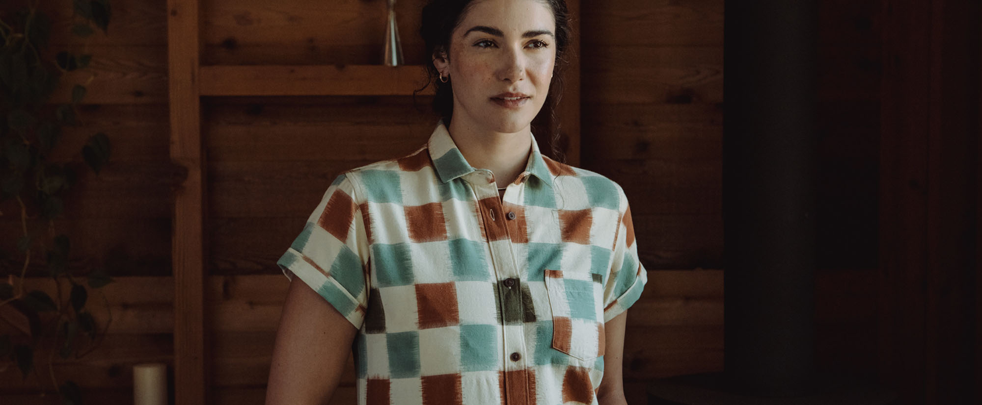 A person with dark hair wearing a short-sleeve, checkered shirt in blue, white, and brown stands indoors against a wooden wall, looking slightly to the side.