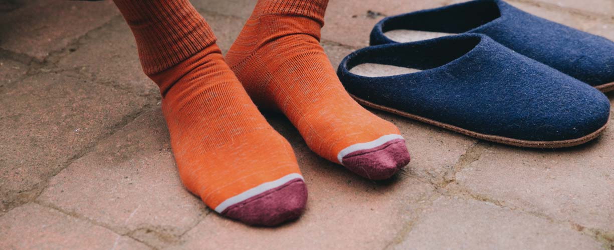 A person wearing orange socks with maroon and gray tips sits on a paved surface next to a pair of navy blue slippers.