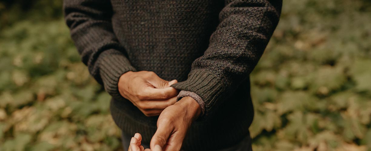 A person in a dark sweater adjusts their sleeve while gently holding a childs hand outdoors, with green foliage blurred in the background.
