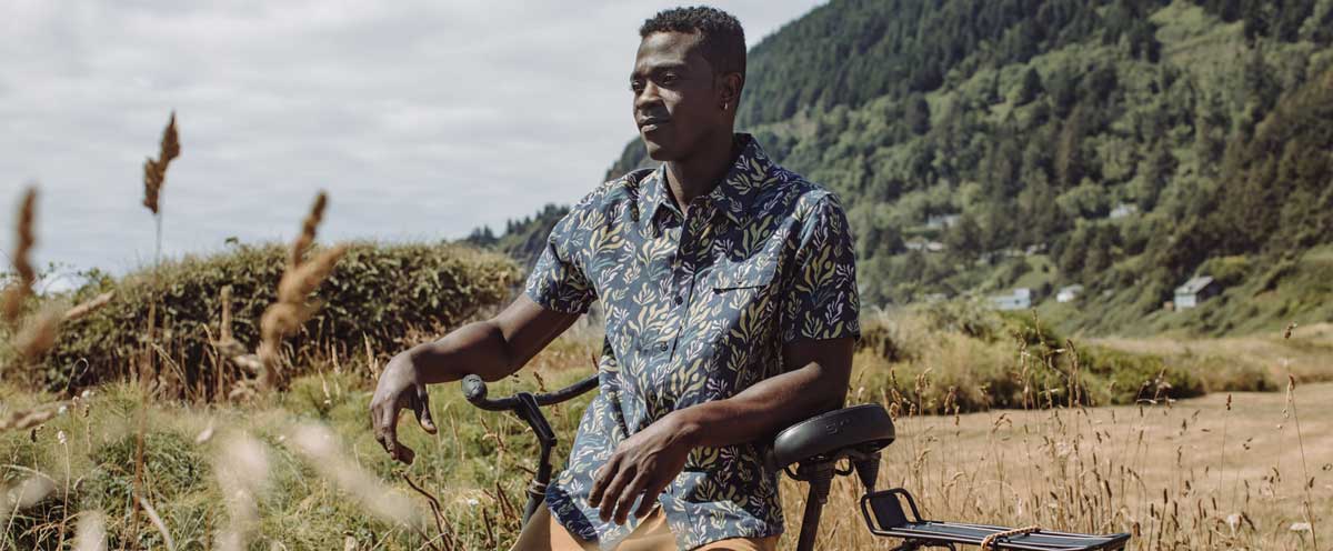 A man in a patterned short-sleeve shirt sits on a bicycle in a grassy field with forested hills and houses in the background under a cloudy sky.