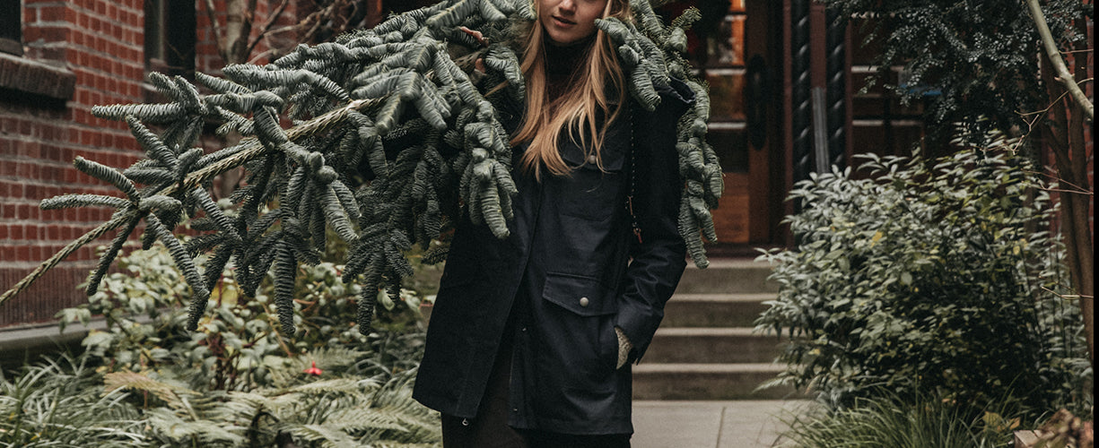 A person wearing a dark coat carries a large, cut evergreen tree over their shoulder while standing in a garden courtyard in front of a brick building.