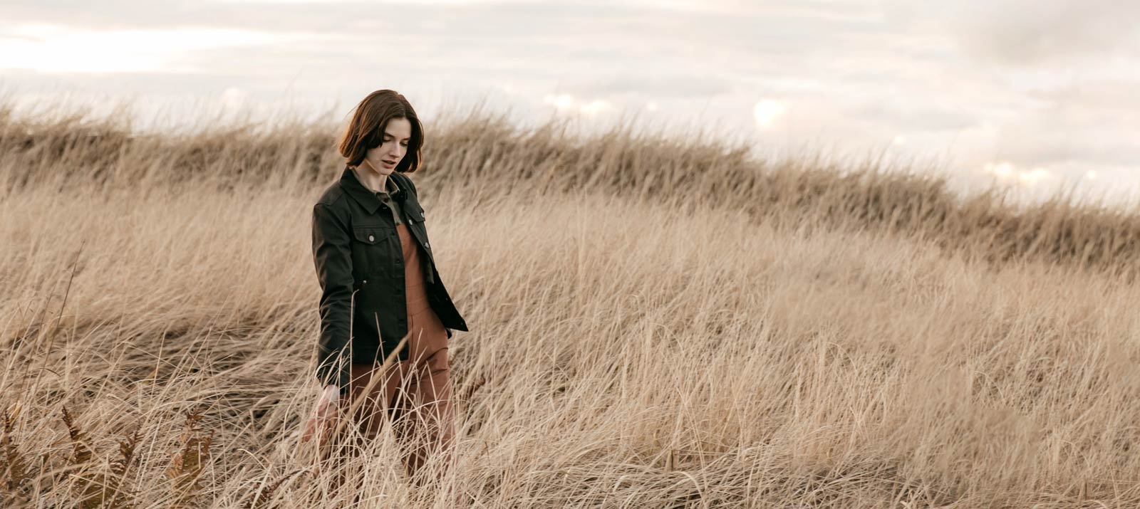A woman with short brown hair, wearing a dark jacket and brown dress, walks thoughtfully through tall, dry grass in an open field under a cloudy sky.