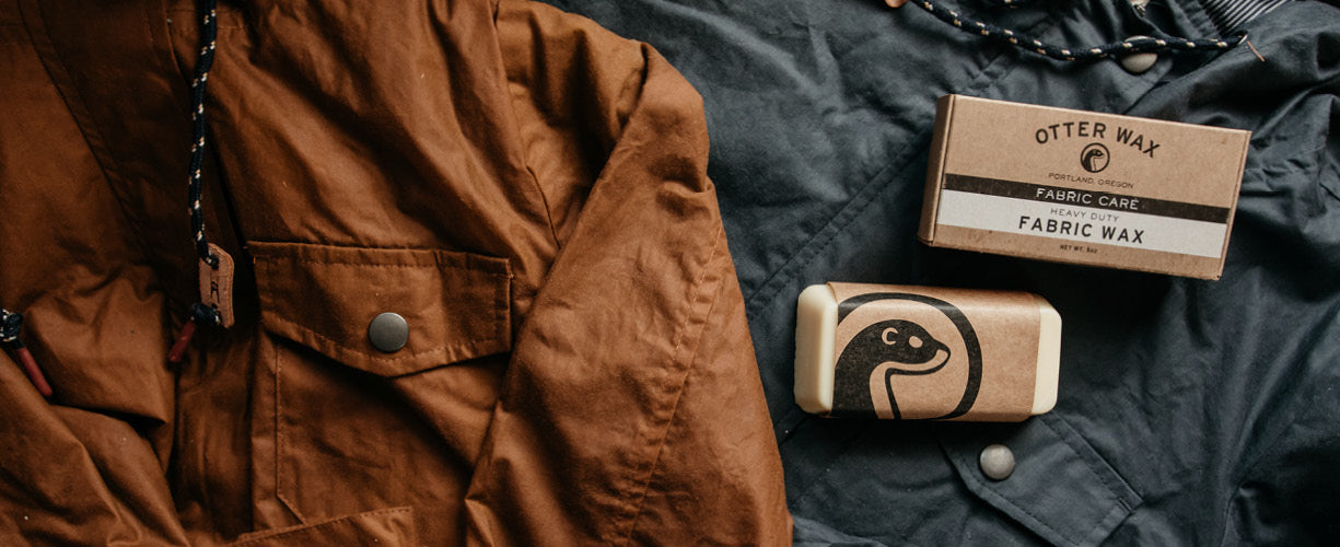 A brown jacket lies next to a bar of Otter Wax fabric wax with an otter illustration and a labeled box, all placed on dark fabric.