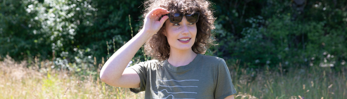 A woman with curly hair and a green T-shirt stands outdoors in a sunny, grassy area, holding sunglasses above her eyes and smiling. Lush green trees are in the background.
