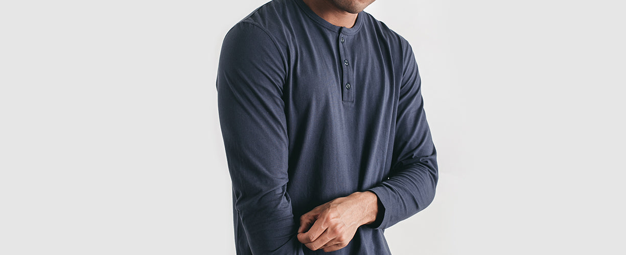 A man wearing a long-sleeve, dark blue henley shirt stands against a plain light background, adjusting one of his sleeves. His face is partially out of the frame.