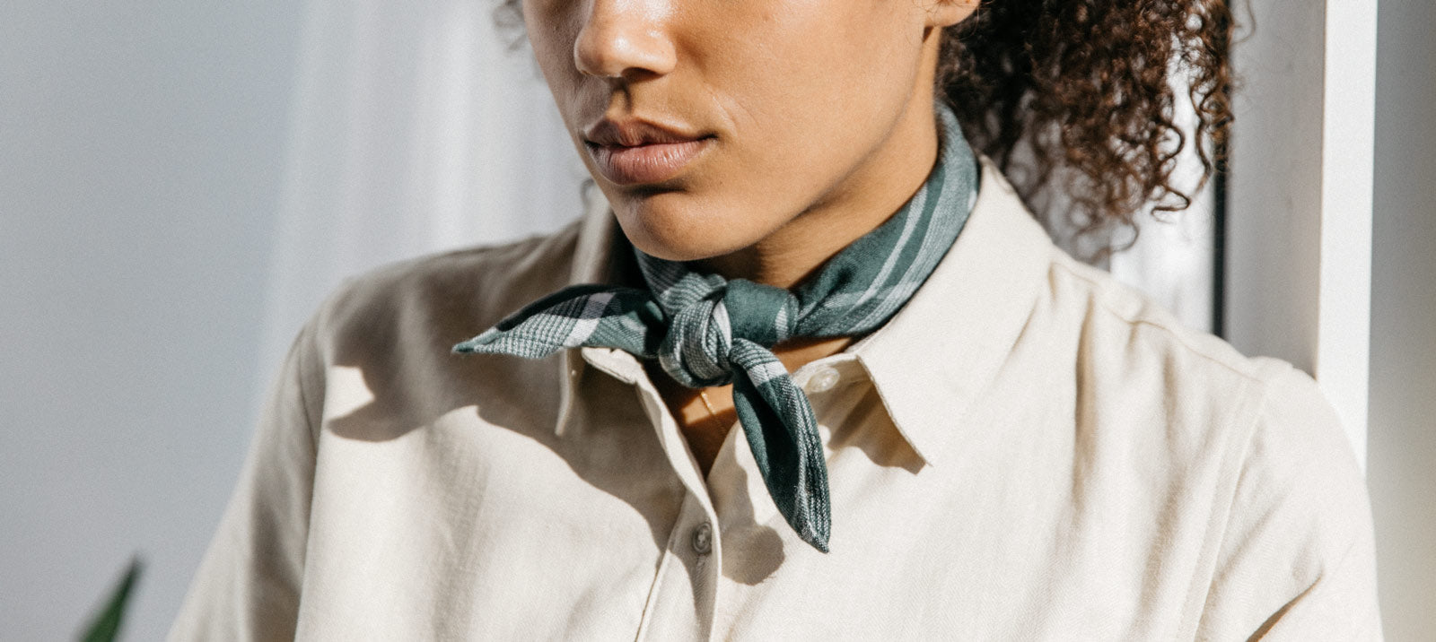 A person wearing a light beige collared shirt and a dark green patterned neck scarf, standing indoors with sunlight casting soft shadows.