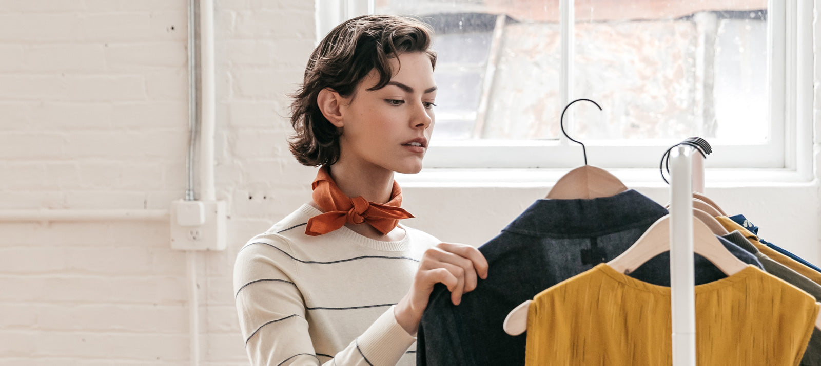 A woman with short brown hair and an orange scarf examines clothes hanging on a rack in a bright room with white brick walls and a large window.