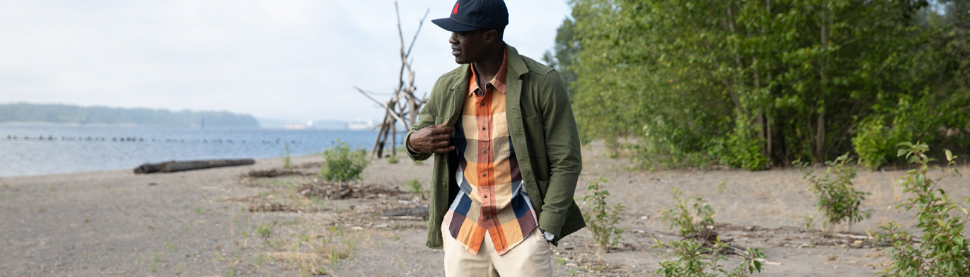A man wearing a baseball cap, green jacket, plaid shirt, and light pants walks along a sandy beach with trees and water in the background.