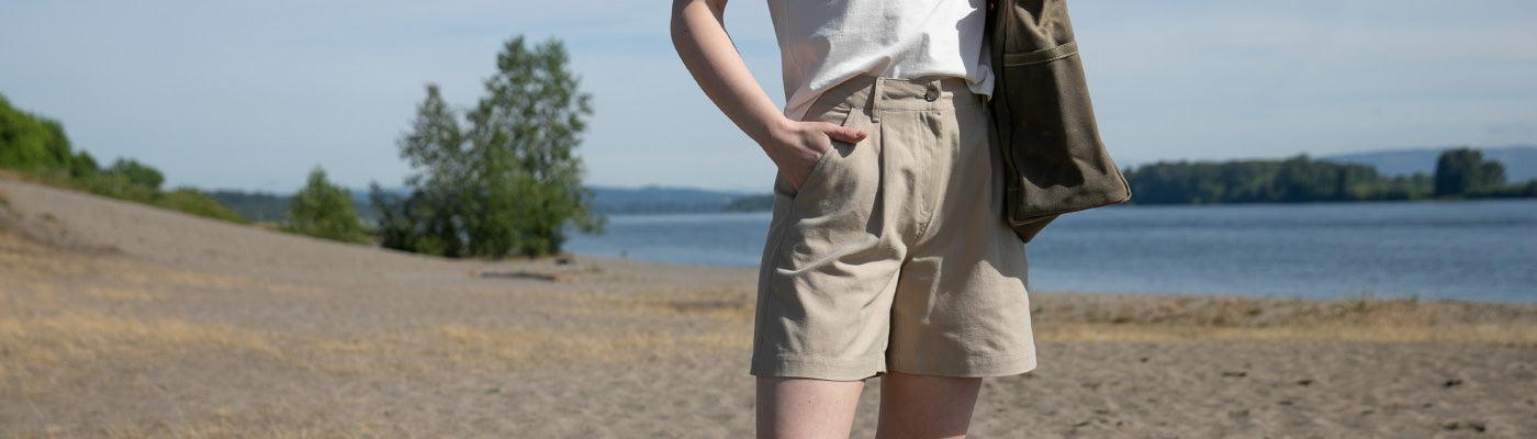 A person stands on a sandy beach near a body of water, wearing beige shorts, a white shirt, and carrying a bag. Trees and distant hills are visible under a clear sky.