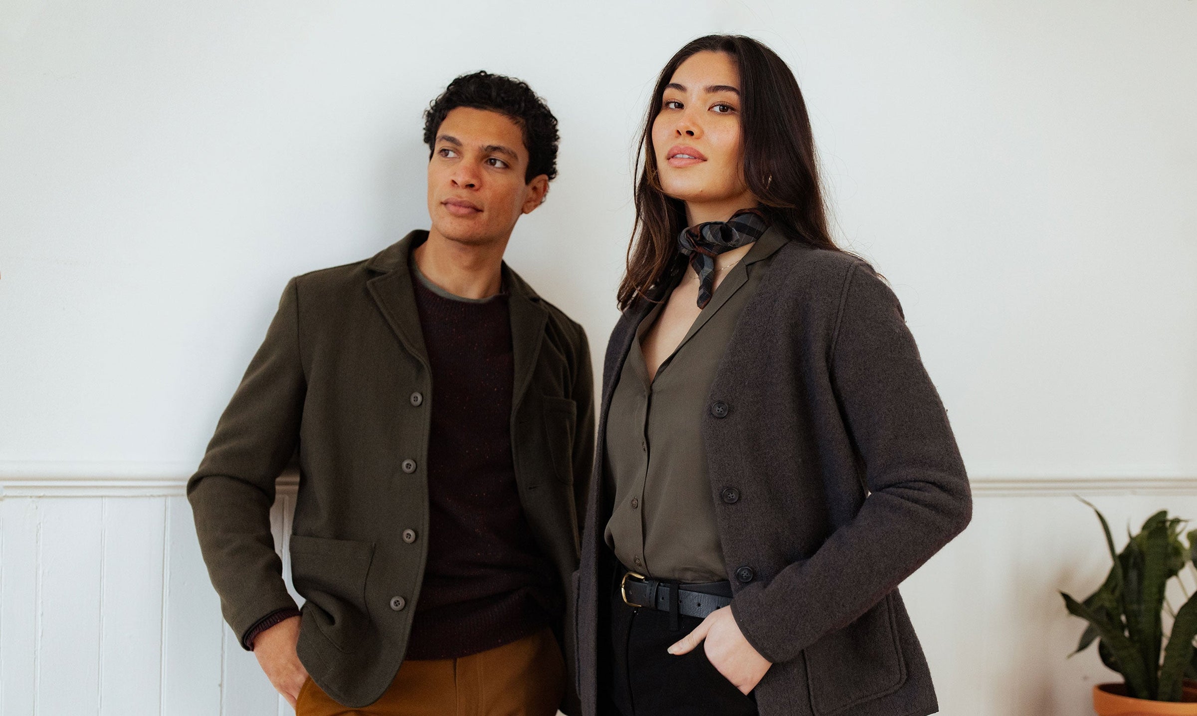 A man and a woman stand confidently indoors, both wearing dark blazers. The woman has a scarf around her neck and one hand in her pocket; a potted plant is visible in the background.