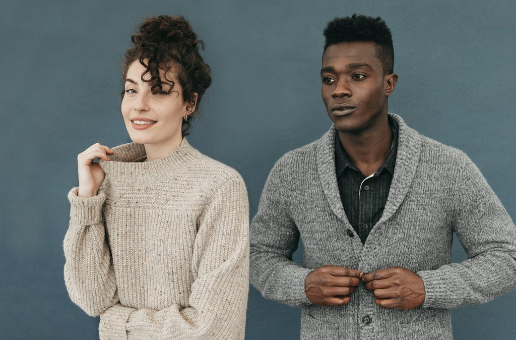A woman in a beige sweater and a man in a gray cardigan stand side by side against a blue background, both looking slightly off camera with neutral expressions.