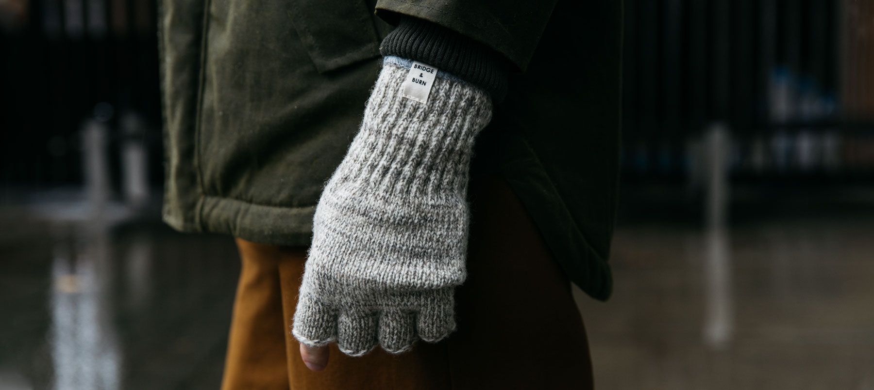 A person wearing a green jacket and brown pants stands outdoors, showing a close-up of their hand in gray wool fingerless gloves on a rainy day.