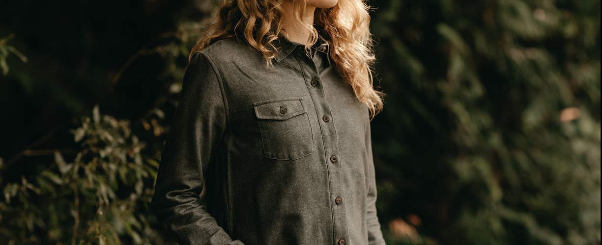 A person with wavy blonde hair wears a button-up olive green shirt with a chest pocket, standing outdoors in front of leafy greenery.