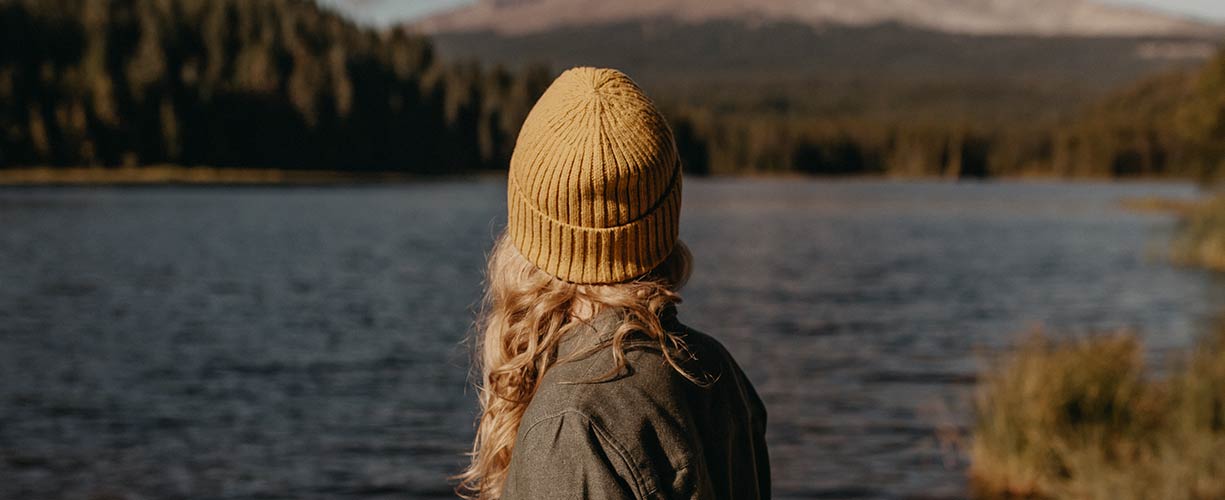 A person with long blonde hair, wearing a yellow knit beanie and a dark jacket, stands by a calm lake with a forest and mountains in the background.