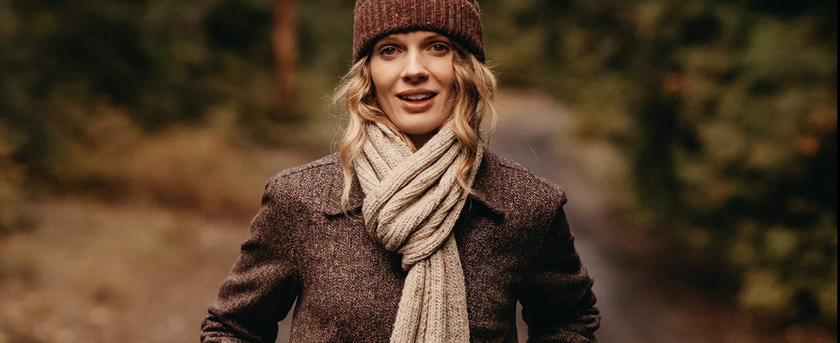 A woman wearing a brown knit hat, beige scarf, and brown coat stands outdoors on a forest path, smiling slightly with trees and foliage in the blurred background.
