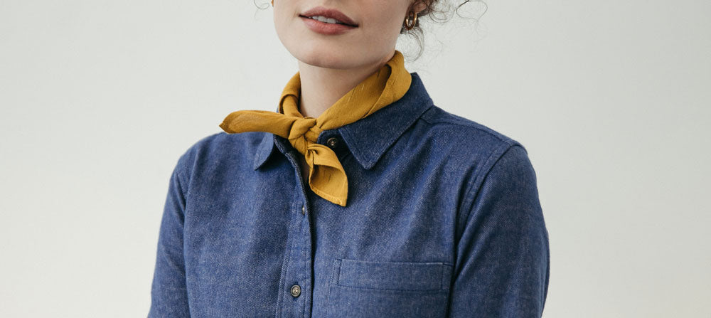 A woman wears a blue collared shirt and a yellow scarf tied around her neck. Only the lower half of her face and upper torso are visible against a plain background.