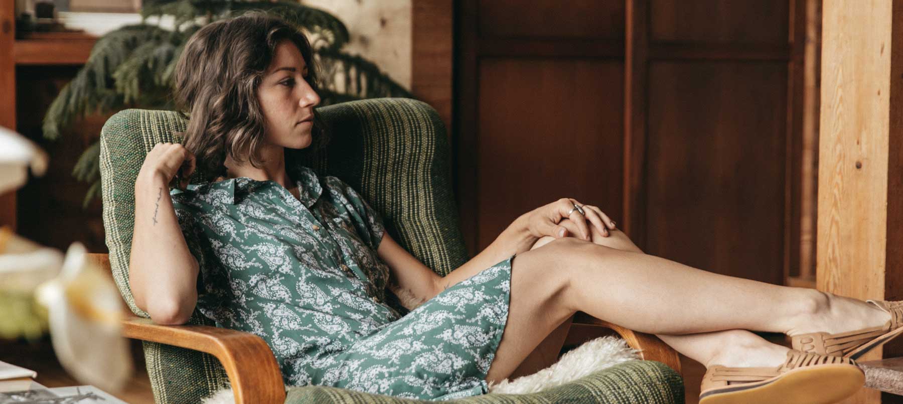 A woman in a green patterned dress sits in an armchair with her legs stretched out, looking to the side with a thoughtful expression in a cozy, wood-paneled room.