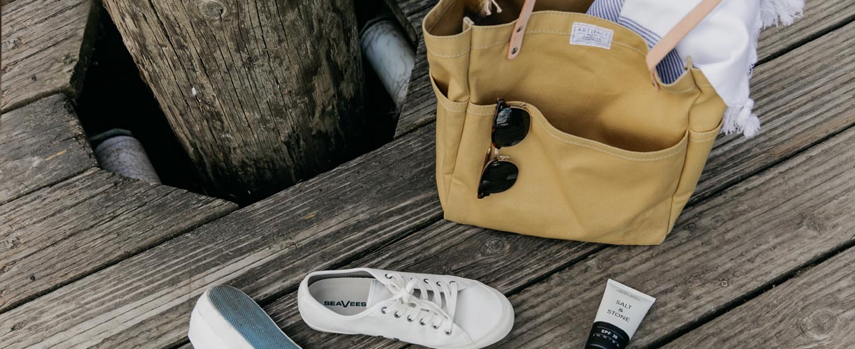 A tan tote bag with sunglasses and a towel inside sits on a wooden dock next to white sneakers and a tube of sunscreen.