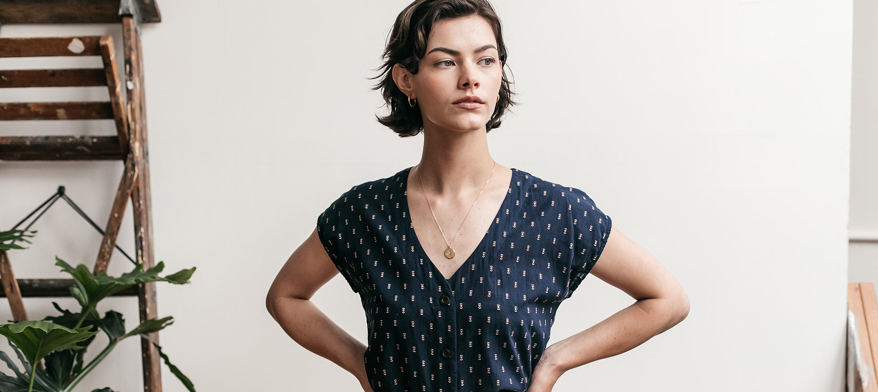 A woman with short brown hair wearing a navy blue patterned dress stands indoors with her hands on her hips, looking confidently to the side. A wooden ladder and potted plant are in the background.