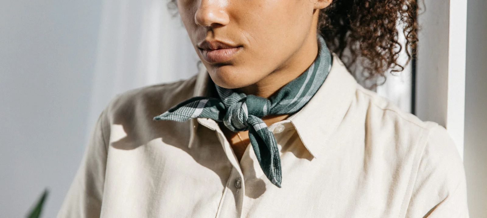A person wearing a light-colored button-up shirt and a green scarf tied around their neck, with curly hair visible, stands indoors in soft natural light.