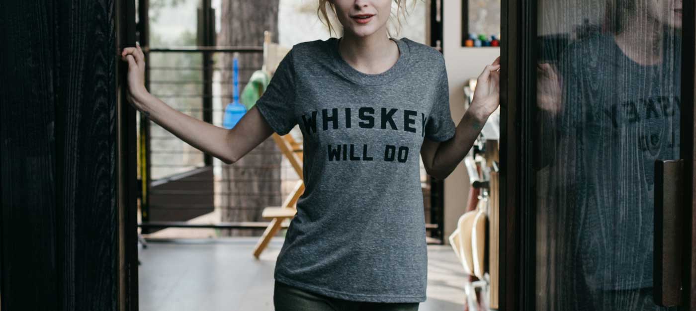 A person stands in a doorway, wearing a gray T-shirt that reads WHISKEY WILL DO. The background shows a modern, sunlit interior with large windows and wooden elements.