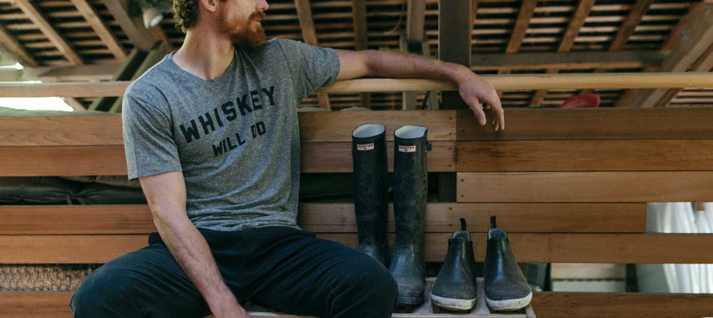 A man with a beard sits on a wooden bench in a barn, wearing a gray WHISKEY MILLED t-shirt and black pants, with pairs of black rubber boots lined up beside him.