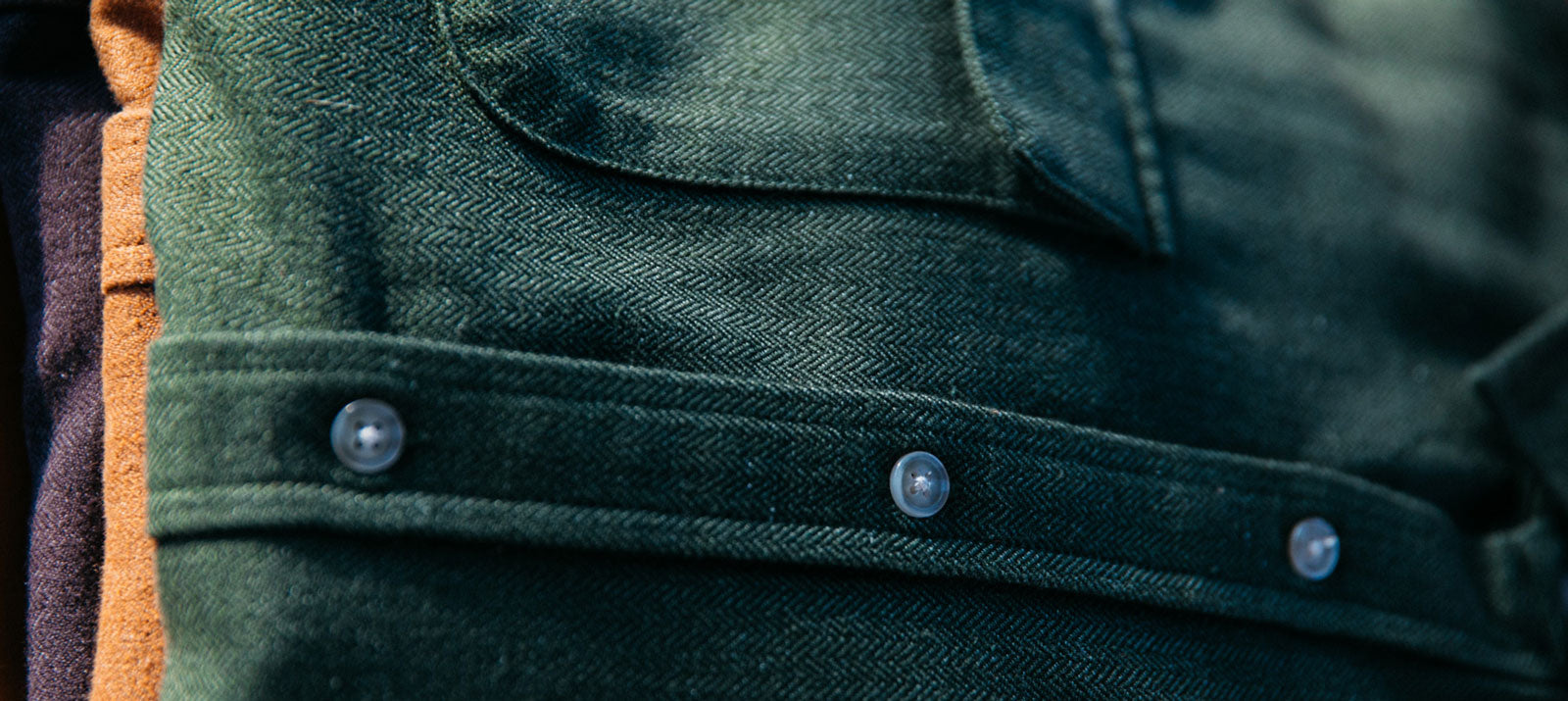 Close-up of green herringbone-patterned fabric with a flap, metal snap buttons, and a pocket, showing detailed texture and stitching. Other colored fabrics are partially visible on the left.