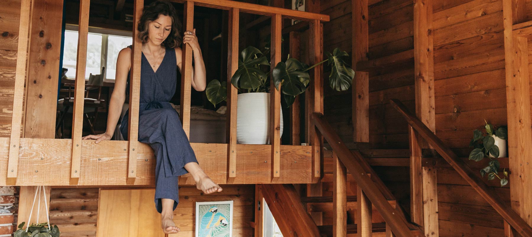 A woman in a blue jumpsuit sits on a wooden railing inside a cozy, wooden house with potted plants and natural light streaming in.