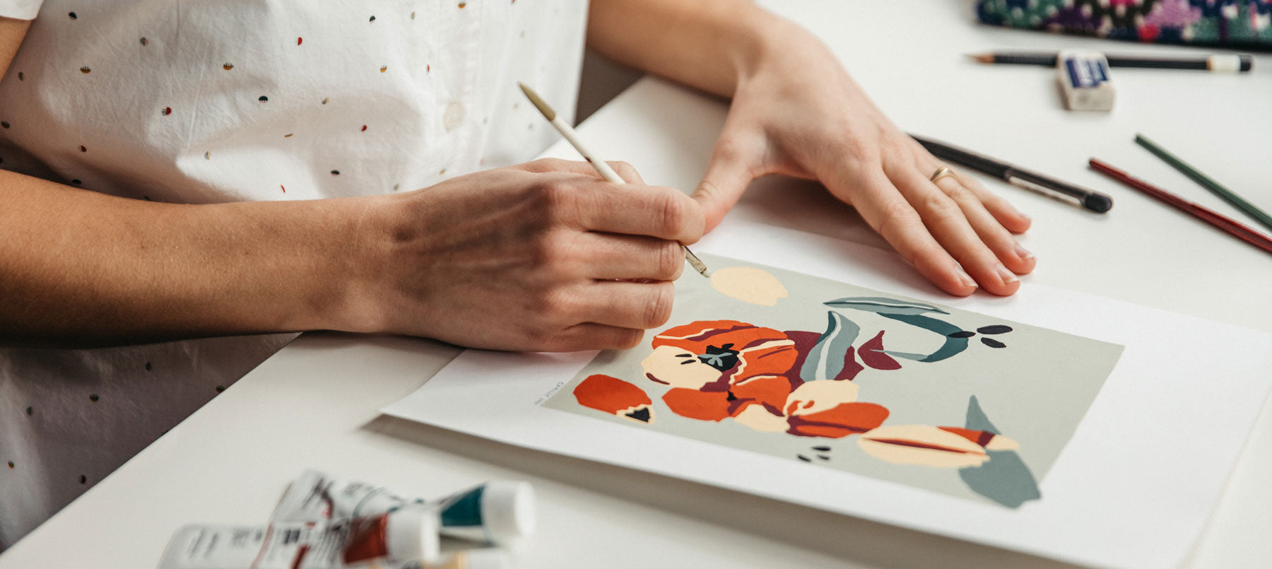A person in a white, polka-dotted shirt paints a floral design on paper at a desk, surrounded by paint tubes, pencils, and erasers.