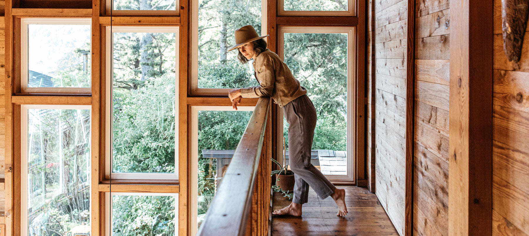 A person wearing a hat leans on a wooden railing inside a cozy cabin with large windows, looking outside at lush green trees.