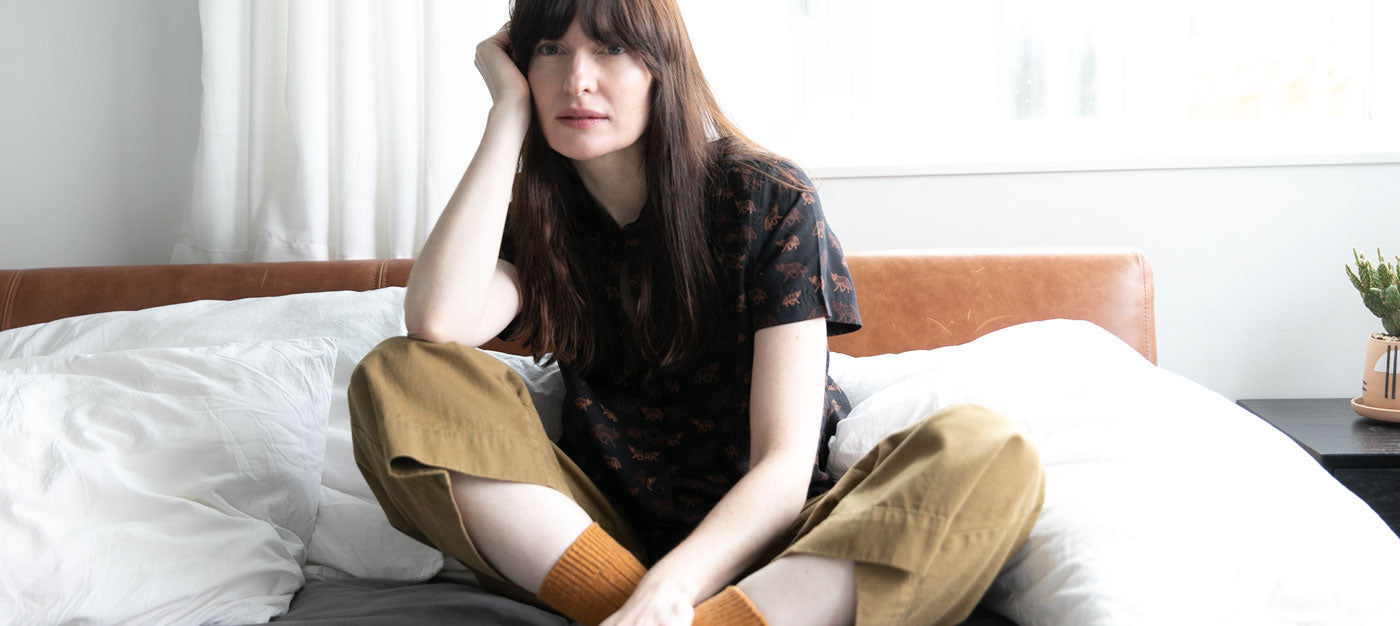 A woman with long brown hair sits cross-legged on a bed, resting her head on her hand. She wears a dark patterned shirt, tan pants, and orange socks in a bright, softly lit bedroom.