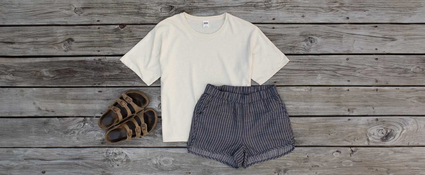 A cream-colored T-shirt, gray striped shorts, and brown sandals laid out flat on a weathered wooden surface.