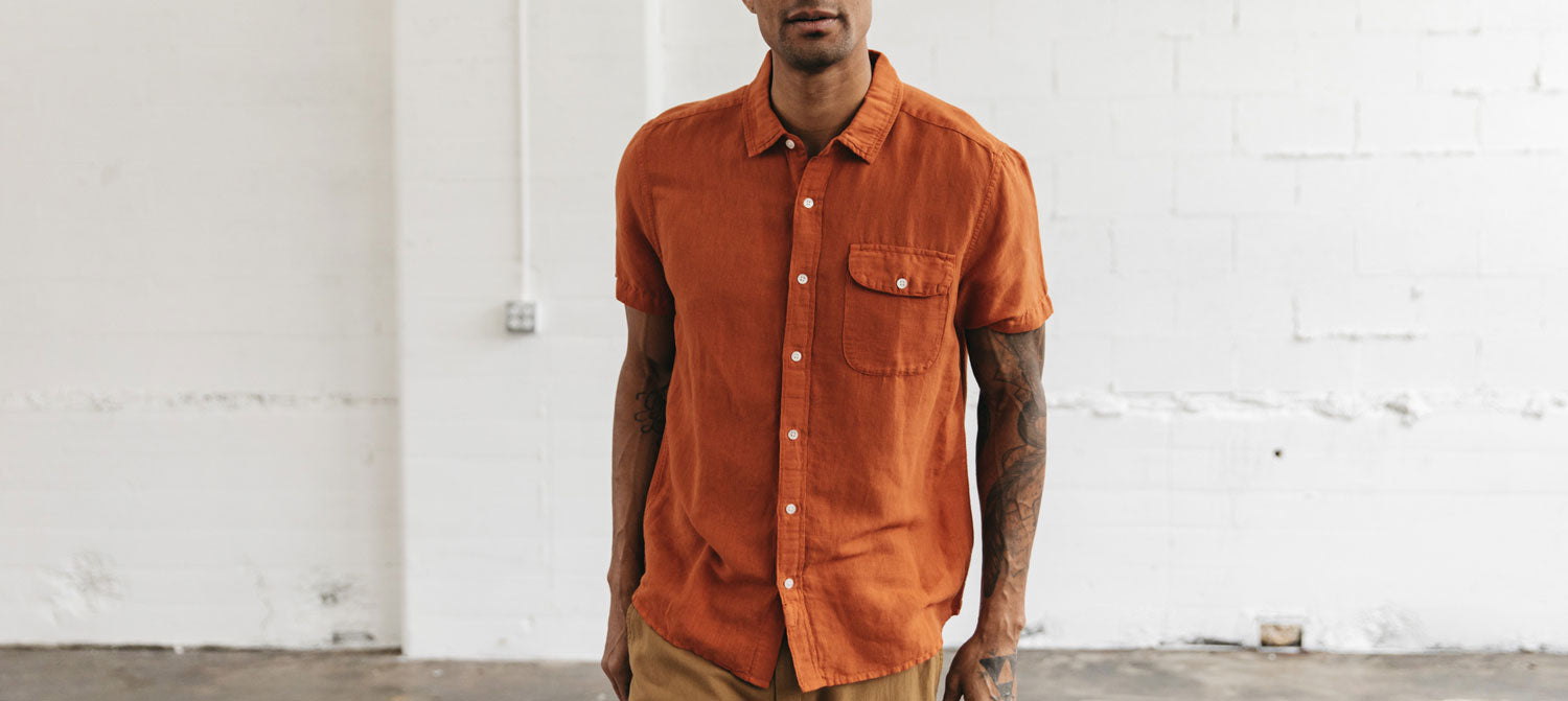 A person wearing a short-sleeved, orange button-up shirt and tan pants stands in front of a white, textured wall in a minimal indoor space.