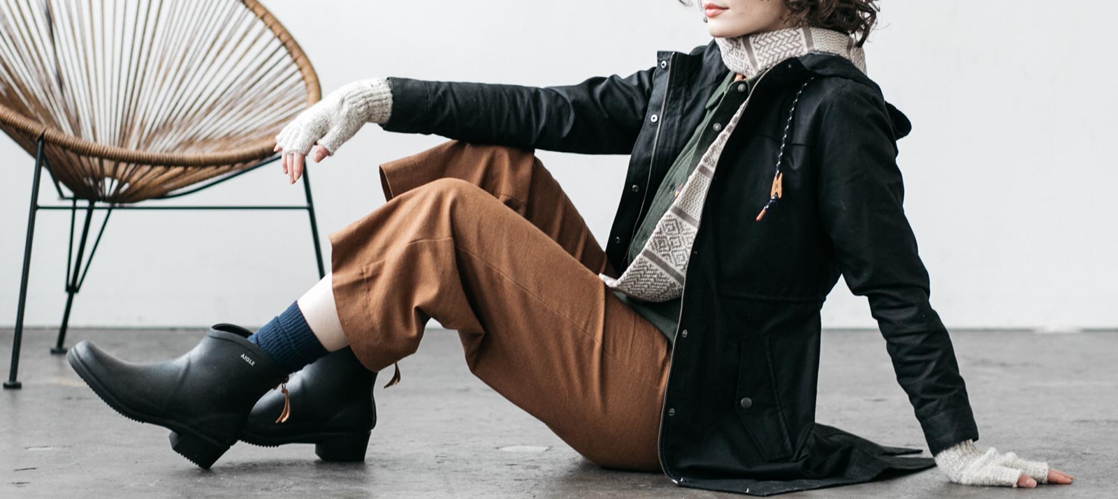 A person in a black jacket, brown pants, and black boots sits on the floor with one arm resting on their knee. A modern chair is in the background. The person wears fingerless gloves and a patterned sweater.