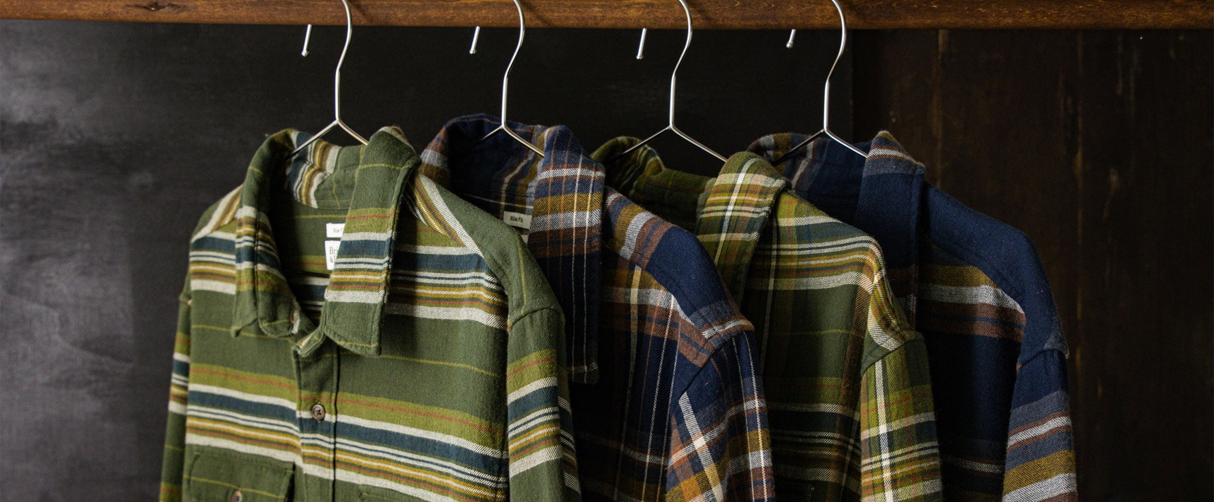 Four plaid button-up shirts in green and blue tones hang on white hangers from a wooden rod against a dark background.