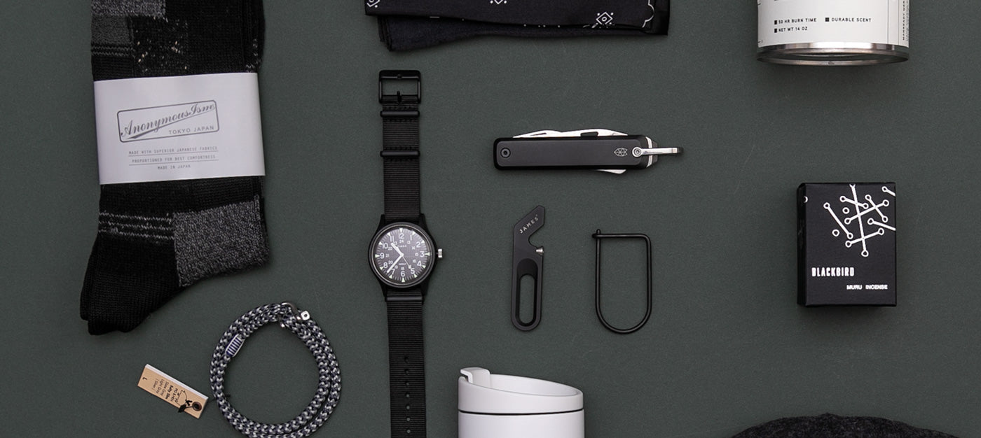 Flat lay of mens accessories on a dark surface, including a watch, socks, black multi-tool, black mug, candle, bandana, rope bracelet, and a box labeled blackout.